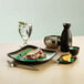 A black Libbey stoneware plate with square detailing on a table with food and a glass of water.