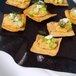 A black GET Stone-Mel display tray with crackers topped with cucumber and hummus and cucumber and peanut spread.