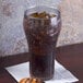 A clear plastic pebbled soda glass filled with dark soda and ice on a table with a napkin and food.