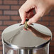 A person's hand opening a Vollrath stainless steel lid with a brass handle.