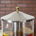 A Vollrath stainless steel lid with a brass handle on a glass container filled with liquid.