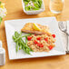 A plate of food with a lemon wedge on top, a bowl of salad, and a glass of water on an Acopa Bright White Square Porcelain Plate on a table.