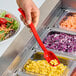 A hand holding a red Cambro perforated salad bar spoon over a bowl of salad.
