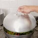 A hand holding a Vollrath metal domed lid over a pan of vegetables.