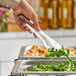 A person holding Carlisle stainless steel serving tongs to a plate of food.