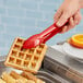 A person holding a Carlisle red plastic utility tong dipping a waffle in red liquid.