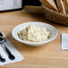 A Tuxton Meridian china bowl filled with mashed potatoes on a table with a basket of bread.