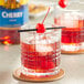 A glass of red liquid with a cherry on top and a toothpick in it.