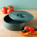 A green round polyethylene container with a lid open next to tomatoes and peppers.