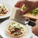 A person uses a Tablecraft stainless steel coarse grater with a black handle to grate cheese on a plate of food.