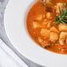 A close up of a white porcelain soup bowl filled with soup, potatoes, and herbs.