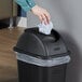 A hand throwing white paper into a black Rubbermaid wastebasket.