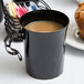 A black WNA Comet disposable plastic coffee cup filled with brown liquid on a table with a muffin.