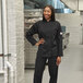 A woman in a black Mercer Culinary Millennia chef's coat stands in a professional kitchen.