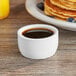 A white cup of syrup on a table next to a plate of pancakes.