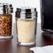 An American Metalcraft clear glass cheese shaker with a stainless steel lid on a table.