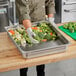 A person in a kitchen with a gloved hand holding a tray of broccoli placing it in a Vollrath stainless steel steam table pan.