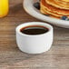 A white ramekin of syrup next to a plate of pancakes on a table.