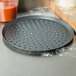 An American Metalcraft round black pan with holes used to make pizza on a counter next to a cutting board and red sauce.