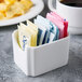 A white container with Libbey bone china sugar packet holders on a hotel buffet counter next to a cup of coffee.