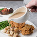 A person pouring gravy from a Reserve by Libbey bone china sauce boat onto a plate of food.