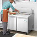 A man in an apron is preparing food on a stainless steel sandwich prep table.