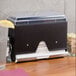 A black Vollrath Straw Boss dispenser on a counter next to a glass of lemonade.