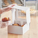An 8 x 8 x 4 inch white bakery box with a clear window, being filled with muffins by a person wearing gloves.