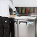 An Avantco 2 door refrigerator with a man in a white shirt and black pants preparing sandwiches on a counter.