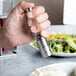 A hand holding a Fletchers' Mill stainless steel and acrylic pepper grinder over a plate of food.