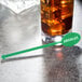 A glass of liquid with ice cubes and a green WNA Comet oval stirrer on a table.