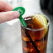 A hand holding a WNA Comet green oval stirrer in a glass of brown liquid with a lime wedge.