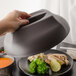 A hand holds a Cambro smoked metal lid over a plate of food with broccoli and meat.