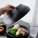 A hand using a black Cambro plate cover to keep food warm on a table.