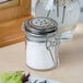 A Tablecraft glass salt shaker with a stainless steel clip-top lid on a table.