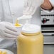 A gloved hand using a Carlisle condiment pump to pour mustard into a container.