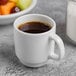 A white Libbey porcelain espresso cup filled with brown liquid sitting next to a bowl of fruit.
