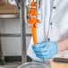 A person wearing blue gloves using a Tellier manual large carrot peeler to peel an orange carrot.