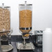 Two Tablecraft stainless steel cereal dispensers filled with cereal on a counter.