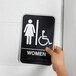 A hand holding a black and white Vollrath Women's restroom sign with Braille.