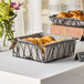 A Clipper Mill black iron square wire basket filled with bread and rolls on a table.