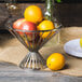 A Clipper Mill chrome plated iron wire basket filled with oranges and apples on a table.