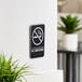 A black and white Vollrath Traex no smoking sign on a wall above a plant.