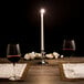 A lit white Hyoola taper candle on a table with two wine glasses.