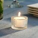 A Leola white votive candle in a glass holder on a table.