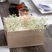 A Leola clear glass wax filled votive candle with a flame on a table with flowers.