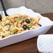 A white rectangular scalloped melamine tray with pasta salad, black olives, and sauce on a table with two spoons.