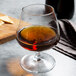 An Arcoroc brandy glass filled with brown liquid on a table.