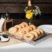 A Clipper Mill clear plastic basket liner with a bagel in a basket on a table with a tray of bagels.