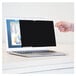 A hand holding a black PrivaScreen filter in front of a laptop with a blurry keyboard.